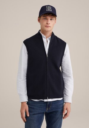 Vesta zip-up de marină, cu design fără mâneci, realizată dintr-un material moale, purtată peste o cămașă albă cu nasturi. Blugii albaștri completează ținuta.