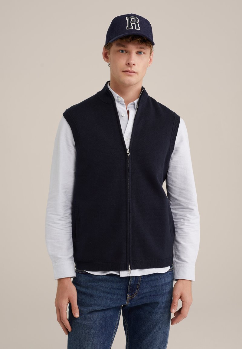 Námořníkova veste s zipem bez rukávů, vyrobená z měkkého materiálu, nošena přes bílou košili s knoflíky. Modré džíny dotvářejí outfit.