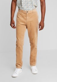 Pantalon en velours côtelé beige avec une texture côtelée, coupe droite, fermeture à boutons et poches, associé à des baskets blanches.
