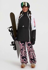 Mlada žena u crno-bijelom skijaškom jaku, u uzorkovanim snježnim hlačama, s rukavicama, kacigom i naočalama za skijanje drži snowboard stojeći uz jednostavnu pozadinu.