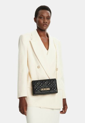 Dunkelhäutige Frau trägt ein cremefarbenes Blazer und einen Rock, dazu eine schwarze, gesteppte Love Moschino Tasche mit goldenem Kettenriemen quer über dem Körper.