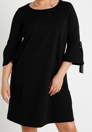 Femme portant une robe noire longueur genoux avec des manches cloche trois-quarts attachées par des rubans noirs, et des boucles d'oreilles pendantes en or.