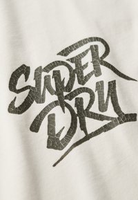 Tela blanca con un gran diseño de texto gráfico negro texturizado que dice "Super Dry" con un detalle de superficie agrietada.