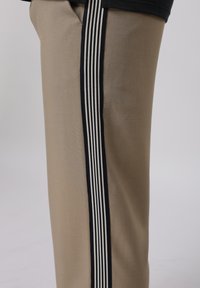 Μπεζ παντελόνι με μαύρη και λευκή κάθετη ριγέ λεπτομέρεια στο πλάι, εμφανιζόμενο από το ισχίο έως το μέσο του μηρού σε απλό φόντο.