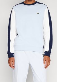 Felpa azzurra con maniche blu navy e bianche, polsini a costine e colletto rotondo. Presenta un piccolo logo verde sul petto.