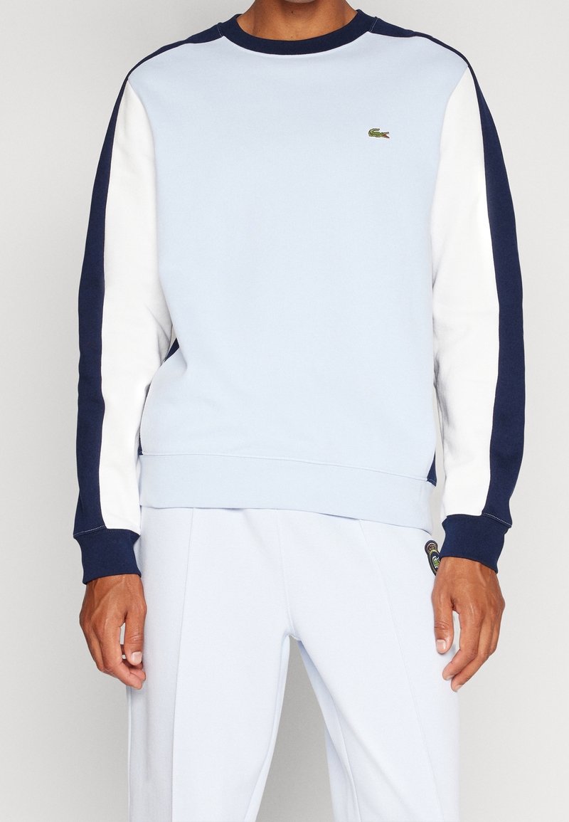 Felpa azzurra con maniche blu navy e bianche, polsini a costine e colletto rotondo. Presenta un piccolo logo verde sul petto.