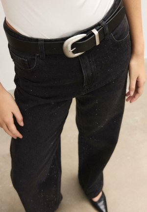 Persona che indossa pantaloni neri scintillanti assicurati con una cintura nera con fibbia argentata, top bianco e scarpe nere con punta, in piedi su un pavimento chiaro.