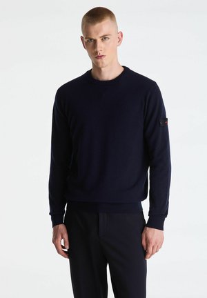 Young man with short hair wearing a dark navy long-sleeve sweater and matching pants, standing against a plain white background.