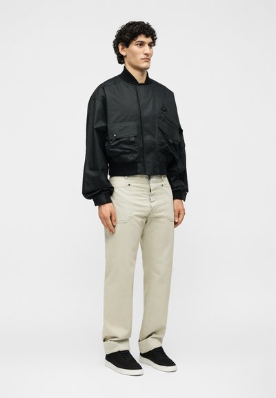 Νεαρός άνδρας στέκεται, φοράει μαύρο μπουφάν bomber με τσέπες cargo, μπεζ παντελόνι και μαύρα αθλητικά παπούτσια, σε ένα μονόχρωμο ανοιχτό γκρίζο φόντο.