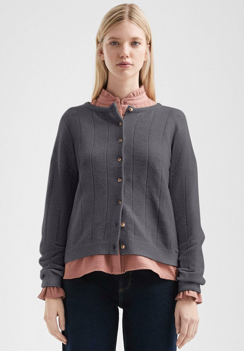 Grijze gebreide cardigan met verticale ribbelstructuur, houten knopen en ronde halslijn. Draag je over een lichtroze gerimpeld shirt, gecombineerd met donkere spijkerbroeken.
