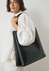 Juodas odinis rankinis krepšys su įmantriu, minimalistiniu dizainu, pasižymintis švelniomis tekstūromis ir erdviu kūnu, laikomas žmogaus, vilkinčio baltą bluzoną.