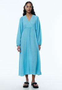 Blaues, strukturiertes Midikleid mit V-Ausschnitt, langen Puffärmeln und geraffter Taille. Kombiniert mit schwarzen Plateausandalen.