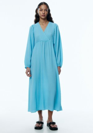 Blauwe, textuur midi-jurk met een v-hals, lange pofmouwen en een gathered taille. Draagt met zwarte platformsandalen.