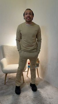 Homme portant un sweat-shirt beige et un pantalon assorti avec des chaussures noires, debout dans une pièce à moquette à côté d'un fauteuil blanc texturé et d'une petite table ronde.