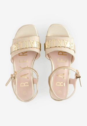 Gold-tone heeled sandals featuring wide straps with the "BAKER" logo, a metallic finish, and an adjustable ankle buckle for fit.