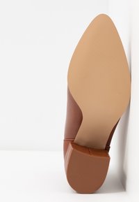 Chaussure à talon haut en cuir marron avec un bout pointu et une texture lisse. La semelle est beige avec un motif de grip légèrement texturé.