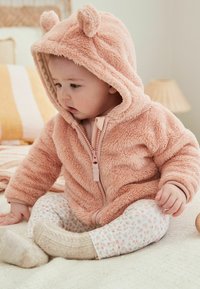 Haut en polaire rose clair avec des oreilles d'ours, fermeture éclair à l'avant, associé à un pantalon blanc avec un petit motif floral ; enfant assis sur une surface texturée.