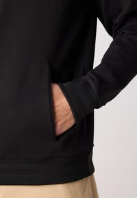 Personne portant une chemise noire à manches longues avec des poignets côtelés, main placée dans la poche avant, pantalon beige visible en bas.