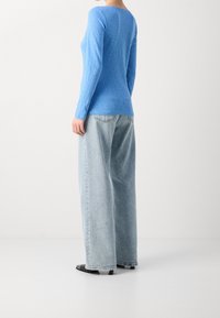 Niebieska bluzka z długim rękawem, wykonana z lekkiej tkaniny, w połączeniu z luźnymi jasnoniebieskimi dżinsami i czarnymi sandałami, widziana od tyłu.