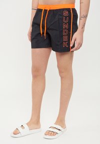 Pantaloni da nuoto neri con coulisse arancione e logo "SUNDEK" sul lato. Realizzati in materiale leggero, con una texture liscia e tasche funzionali.