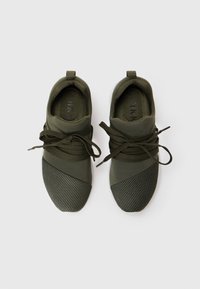 Baskets en olive avec un embout en maille noire texturée, des lacets intégrés et un dessus en tissu lisse, avec une sangle de traction au talon.
