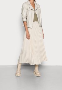 Veste de motard en cuir crème clair, haut olive, jupe midi plissée beige et bottines épaisses crème. Textures lisses et palette de couleurs neutres.