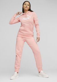 Felpa rosa chiaro con tasca a canguro e logo bianco, abbinata a pantaloni jogger coordinati, con polsini a coste e vestibilità rilassata. Sneakers bianche.