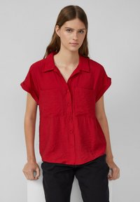 Rotes kurzärmliges Hemd mit Knopfleiste, zwei Brusttaschen, strukturiertem Stoff und einem kragen, kombiniert mit dunklen Hosen.