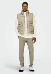 Gilet puffer beige con collo alto e due tasche frontali, indossato sopra un maglione bianco. I pantaloni grigi e le sneakers bianco e nero completano il look.