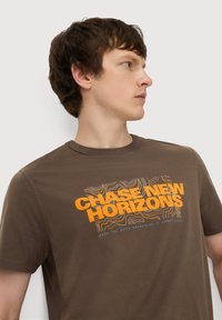 Brun bomulds T-shirt med rund hals, der har et grafisk design med orange og hvid tekst "CHASE NEW HORIZONS" og abstrakte linjer.
