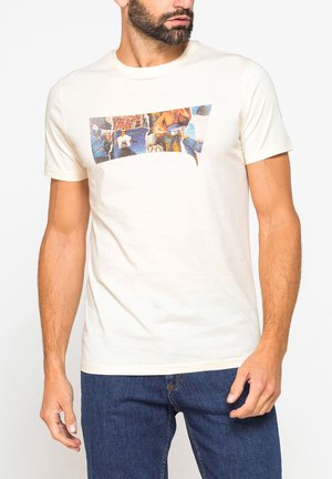 Uomo che indossa una maglietta color crema con un design collage strappato sul petto e jeans blu, in piedi su sfondo bianco.