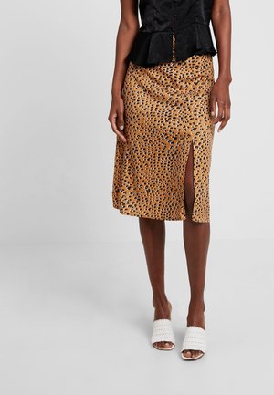 Kvinde iført en tanfarvet midi-nederdel med leopardprint og slids foran, sort tekstureret top og hvide åbne flettede mule-sandaler.