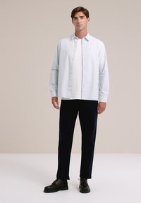 Šviesiai mavi marškiniai su ilgomis rankovėmis virš baltos marškinėlių, derinami su tamsiai mėlynomis kelnėmis ir juodais batais. Švelni medžiaga, laisvas siluetas ir paprastas dizainas.