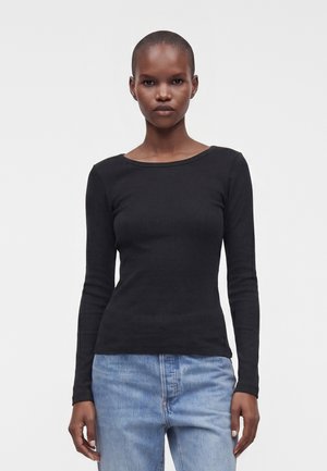 Femme aux cheveux courts portant un haut noir à manches longues moulant et un jean bleu clair, debout devant un fond blanc uni.