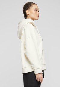 Light beige hooded sweatshirt with a front pocket and star accents on the sleeve. Features adjustable drawstrings and a relaxed fit.