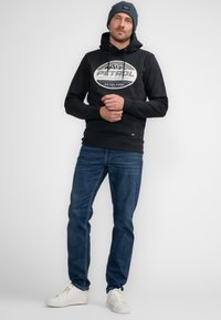 Sudadera negra con un logo gráfico en el frontal, combinada con jeans azules y zapatillas blancas. La sudadera tiene un cordón y un bolsillo canguro.