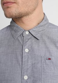 Šedá košile na knoflíky s límečkem, vybavená bílými knoflíky a malou nášivkou loga v červené, bílé a modré barvě na náprsní kapse.