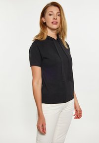 Haut à capuche noir à manches courtes avec un cordon au cou, fabriqué dans un tissu lisse et léger. Associé à un pantalon blanc, en position debout.