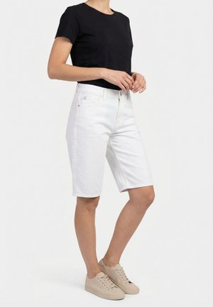 Persoon draagt een zwart T-shirt met korte mouwen, witte knielange spijkershorts en beige sneakers tegen een effen witte achtergrond.