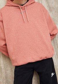 Rosa hoodie med dragsko i huvan och en liten silverlogotyp på bröstet, parat med svarta byxor som har en vit logotyp.