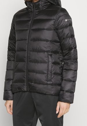 Person iført en sort quiltet hættetrøje med lynlås og Champion-logo på venstre ærme, kombineret med sorte bukser.