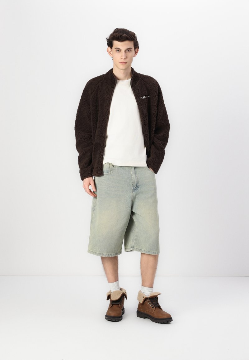 Brązowa polarowa kurtka, biała koszulka, jasnoniebieskie szorty i brązowe buty z beżowymi akcentami. Model stoi na jednolitym tle.