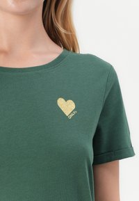 Zelena pamučna majica s kratkim rukavima i zaobljenim izrezom, s zlatnim šljokicama u obliku srca i logom "ONLY" na prsima.