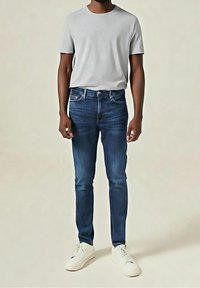 Man som bär en ljusgrå t-shirt, mörkblå slim-fit jeans och vita sneakers, stående mot en enhetlig ljus bakgrund.