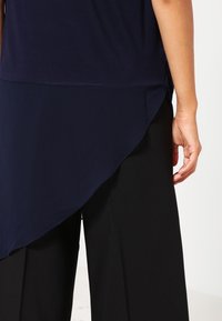 Navy blue, flowing top with a curved hem, layered design; paired with black, wide-leg trousers. Smooth fabric with a subtle sheen.