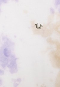 Närbild på ett tyg med lila och beige batikmönster samt en liten metallisk hästskoformad emblem.