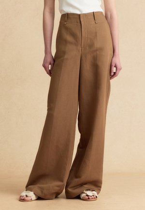 Persona che indossa pantaloni marroni a vita alta e gamba larga e sandali bianchi in maglia con punta aperta, in piedi davanti a uno sfondo beige.