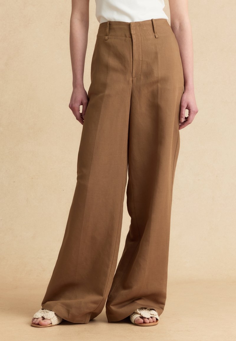 Persona con pantalones marrones de talle alto y pierna ancha y sandalias de punto blancas con los dedos descubiertos, de pie sobre un fondo beige.