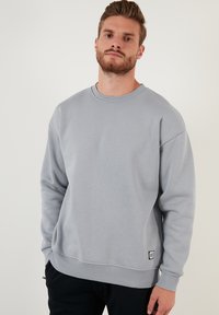 Sudadera de cuello redondo gris con puños y dobladillo acanalados. Tejido suave, corte relajado, con una pequeña etiqueta de la marca en el lado inferior izquierdo.