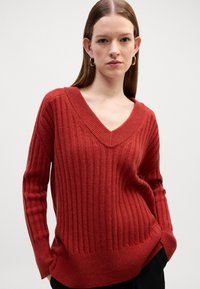 Czerwony sweter z prążkowanej dzianiny z głębokim dekoltem w kształcie litery V, długimi rękawami i luźnym krojem. Materiał wydaje się miękki, z teksturowanym wykończeniem.